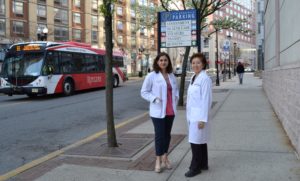 Two women standing on a sidewalk next to a city street wearing physician whitecoats