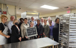 A large group of people standing in a commercial kitchen, with the person in the center holding a sign that reads, ELIJAH'S PROMISE / FOOD CHANGES LIVES