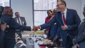 President Masisi and President Holloway smile and shake hands across a conference table surrounded by people.