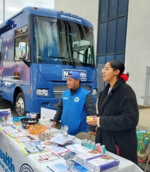 Photo of two people standing at a community health fair table, next to a New Jersey Department of Health van. They are staff members working for Rutgers Global Health Institute and New Jersey Department of Health, and their table is full with materials such as pamphlets, testing kits, promotional items.