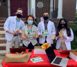 Photo of four people standing outside in front of a building's steps. Three of the people are wearing white medical coats. Two of the people are holding "Veggie Rx" brochures, and some are holding "prescription pads" that are part of the Veggie Rx program to encourage eating healthy foods.