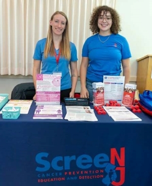 Two people standing behind a table that says Screen NJ on it - Screen NJ is a cancer prevention, education, and detection resource in New Jersey. On the table there are a variety of materials, such as flyers, promotional items.